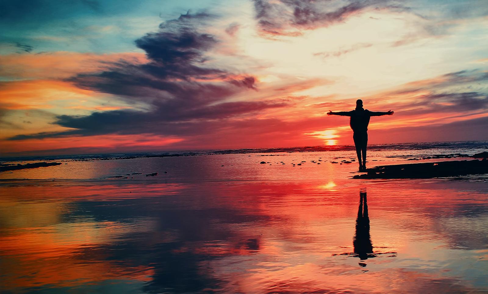 A person standing at the shore at sunset with their arms open wide, the sky is the limit